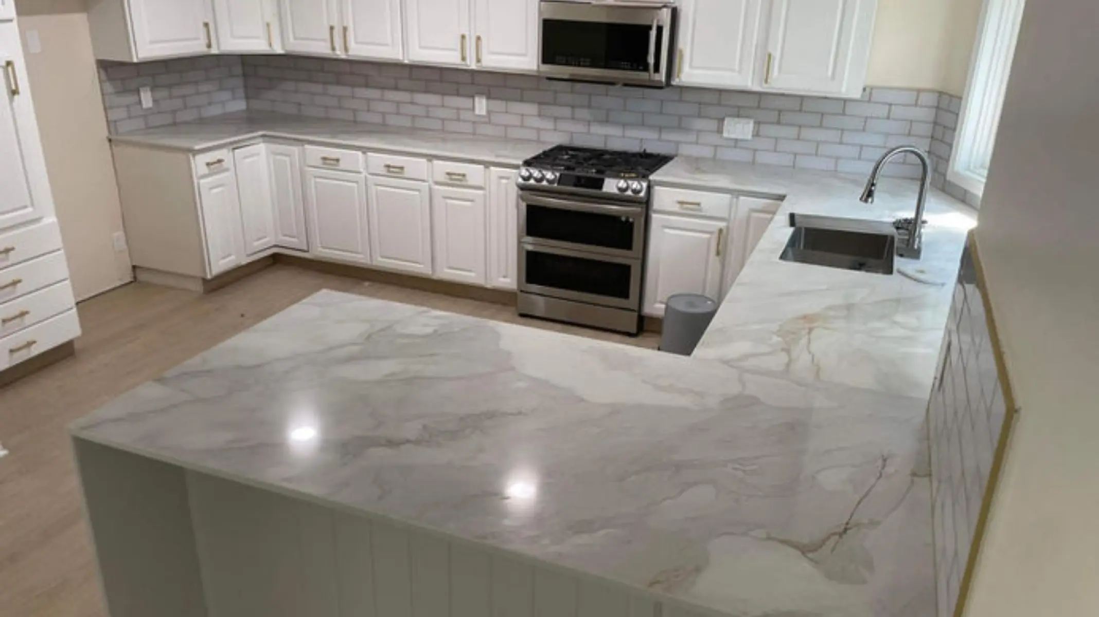 Clean, modern kitchen with white cabinetry, light gray marble countertops, stainless steel stove and microwave, and a stainless steel sink beneath a window.